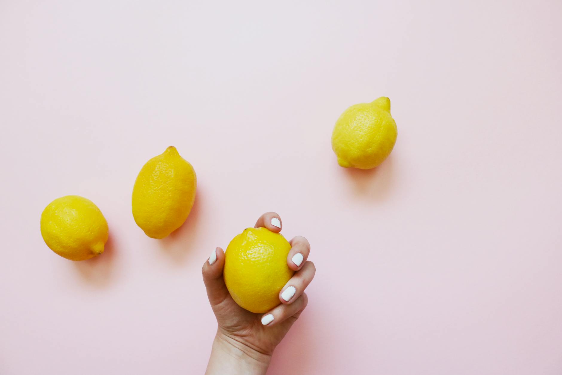 A hand holding a fresh lemon against a soft pink background surrounded by additional lemons, representing natural vitality and sensory pleasure