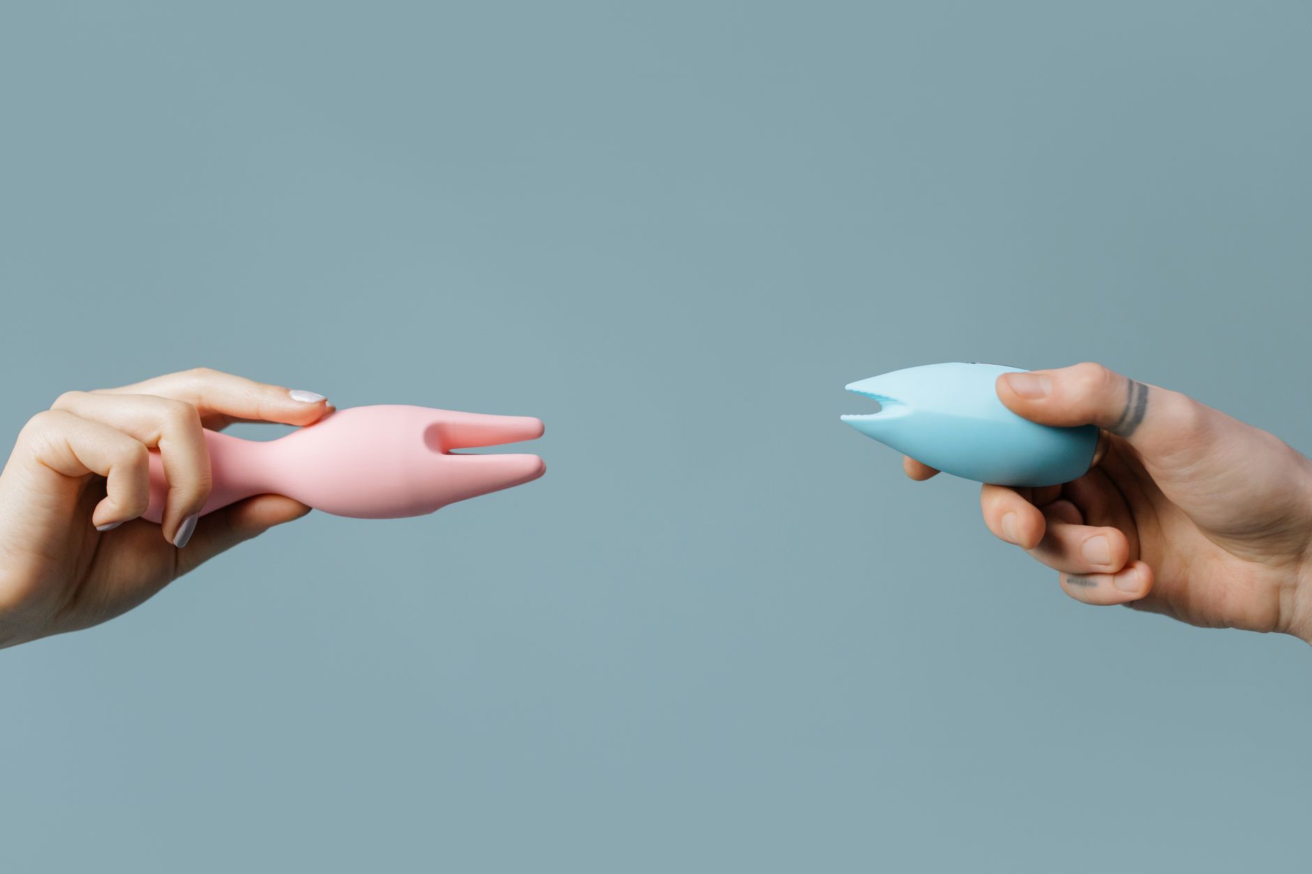 Two hands holding pink and blue silicone clitoral vibrators against a pastel background, symbolizing personal pleasure and choice.